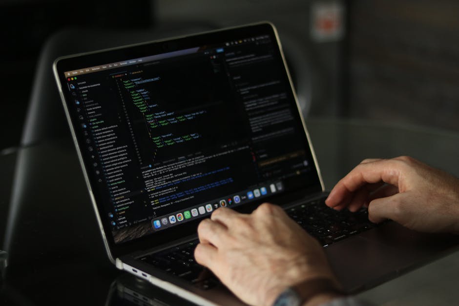 A person using a laptop on a dark surface, the screen displaying lines of code with colorful syntax highlighting, suggesting programming or web development activity. The person's hands are visible, wi